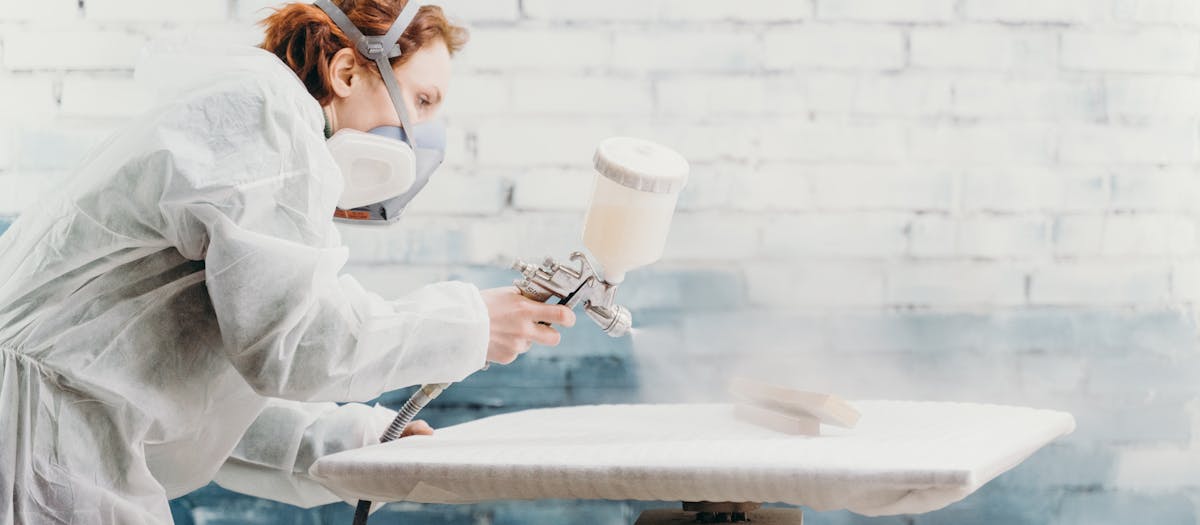 Redheaded woman wearing protective gear spray paints a furniture piece indoors.